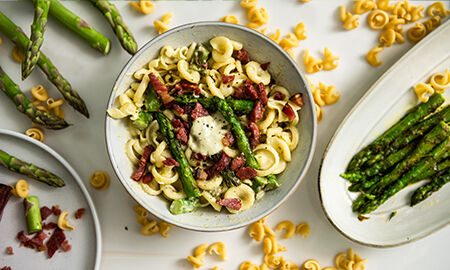 Présentation de la recette de Pâtes, asperges et jambon d'Ardenne sur une table avec les différents ingrédients en décoration dont un plat d'asperges, des pâtes éparpillées et quelques dés de jambon d'ardenne