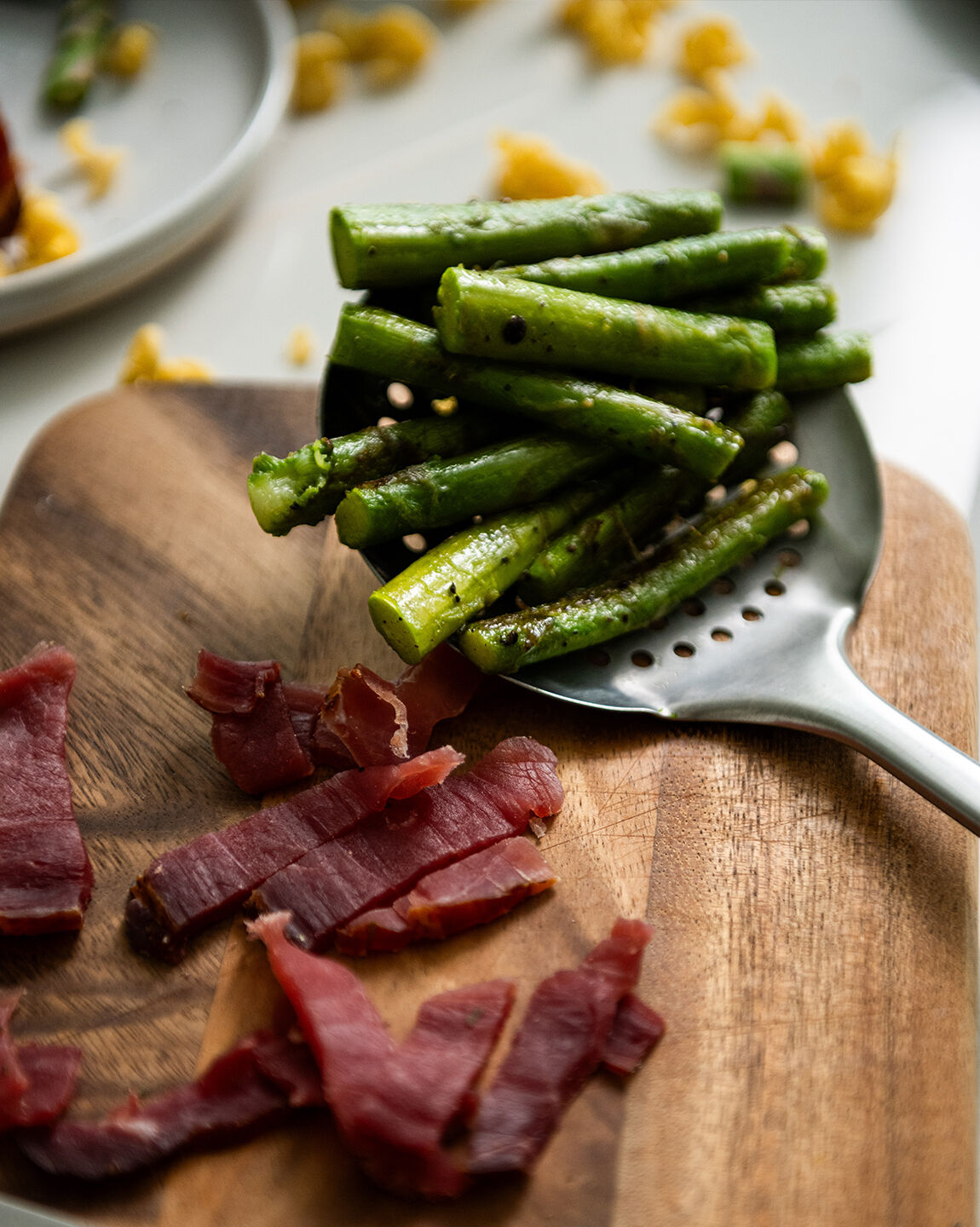 Présentation d'asperges cuites à la poêle dans une cuillère posée sur une planche en bois à côté de morceau de jambon d'ardenne et quelques pâtes en décoration